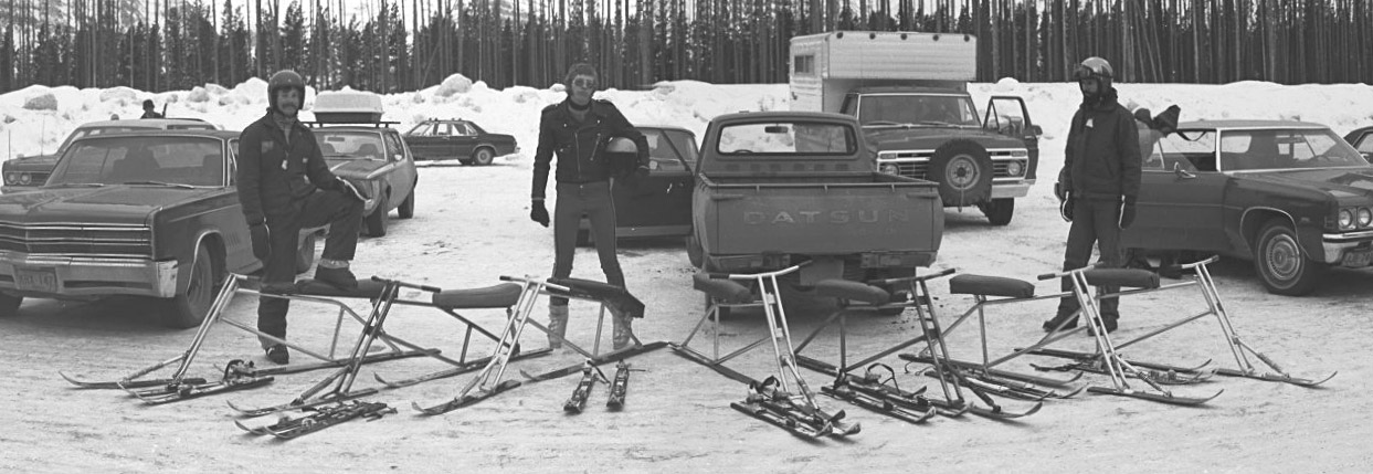 This remarkable image is one of a series uncovered by Dave Agnew. It was taken during the 1975-76 ski season and shows <br />a youthful Dave Agnew, Kevan Leycraft and Ted Kuchura in the Lake Louise parking lot surrounded by some very rare <br />early skibikes. Incredibly, this was also the first season that Kevan's skibikes were ridden at Lake Louise, Alberta.  <br />The bike that Dave has his foot on is the venerable "Red Rocket", described in the SkiBike History page. <br />Only Dave's bike and the one next to it have long-travel shock absorbers under the seat. These may <br />have been the first bikes ever to have incorporated this feature. Note that all of these bikes have their <br />handlebars welded directly to the top of the dual steering columns. Also of interest is the skibike in front of <br />Kevan, which has a hinged seat which wraps around the rear frame member and is supported on two short-travel springs.  <br />The four machines on the right incorporated an early frame geometry and seat design, where the seat was mounted far forward on the <br />frame and supported by two small short-travel springs. With the exception of Dave's skibike, none of the machines in this photo exist today.<br />Thanks again Dave for the great pictures!